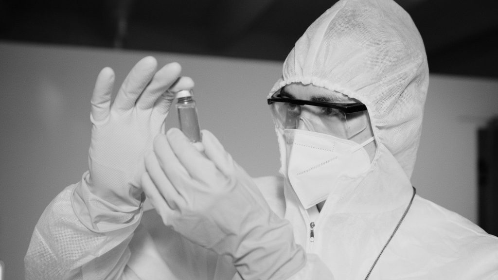 Researcher in protective gear inspecting a vial in a lab setting.